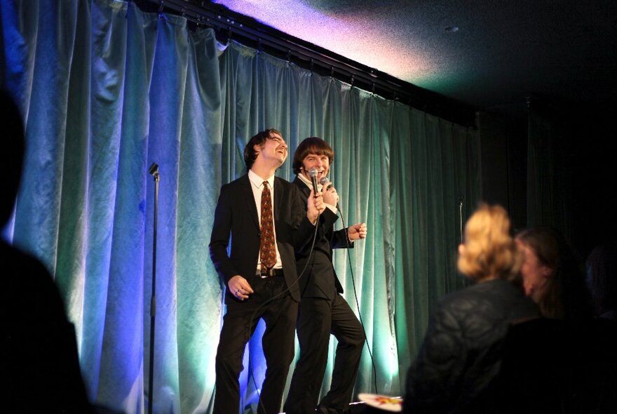 Two men in suits leaning into each other performing on a small stage in front of an audience.