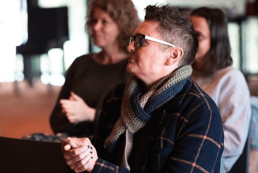 A person with short hair and glasses wearing a tartan jacket and knitted scarf, seated with hands clasped; two women blurred in the background.