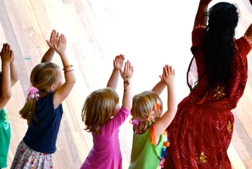 Children dancing with arms raised, standing in a line behind an Indian woman dancing wearing a red sari.