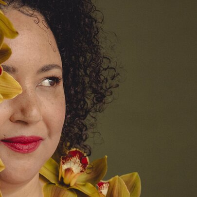A woman with tightly curled hair peeking out from behind some flowers.