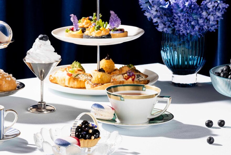Two-tiered plate of savoury and sweet food served as part of a High Tea, with a cup of tea in a saucer and a vase of flowers.