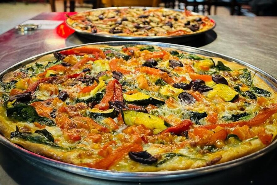 Two large pizzas on a table in metal trays.