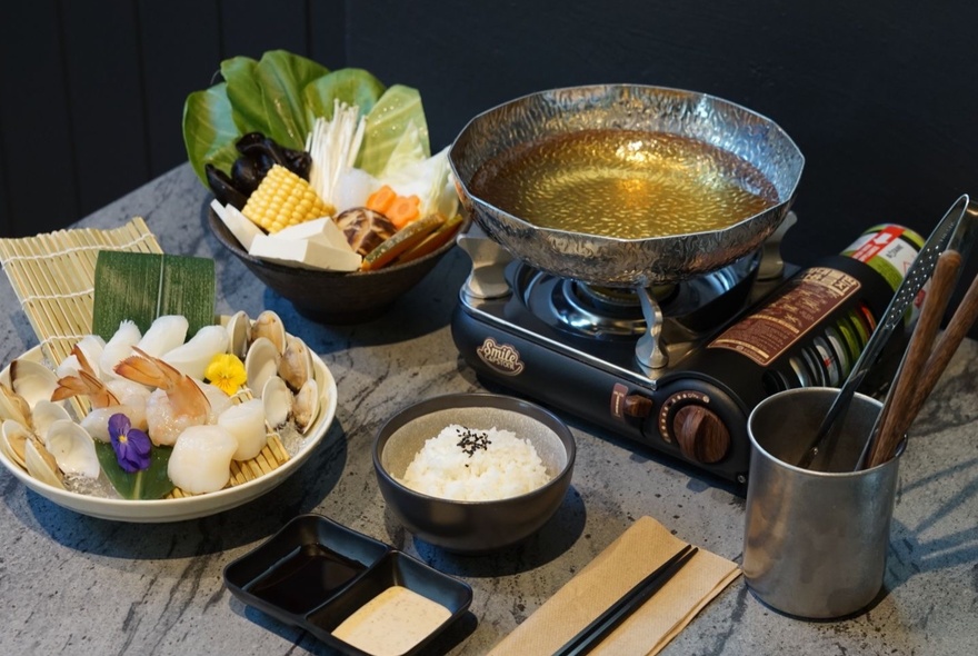 A personal Japanese hot pot set up with seafood and vegetable ingredients prepared in bowls alongside the small tabletop gas burner with stainless steel bowl of broth.