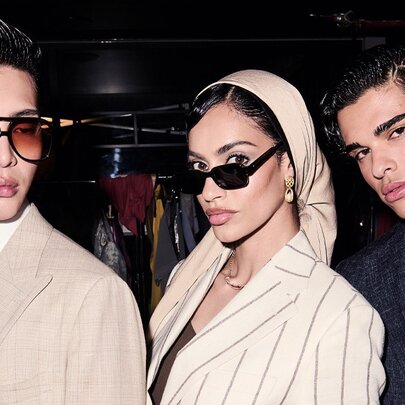 Two fashion models posing backstage in tailored neutral-toned blazers and chic dark sunglasses, with a third fashion model wearing a dark suit.
