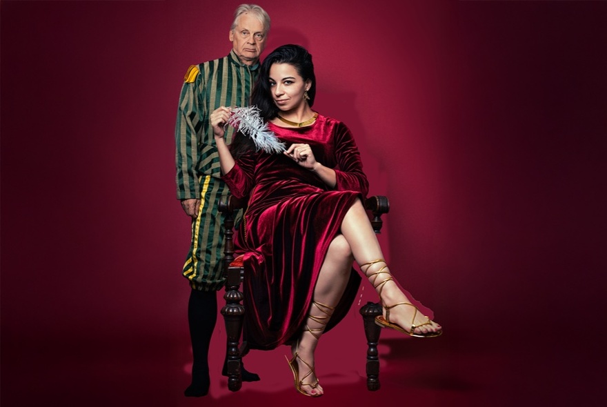 Woman wearing a red velvet dress, seated on a chair with bare legs in sandals crossed, older man in striped knickerbocker suit standing behind her.