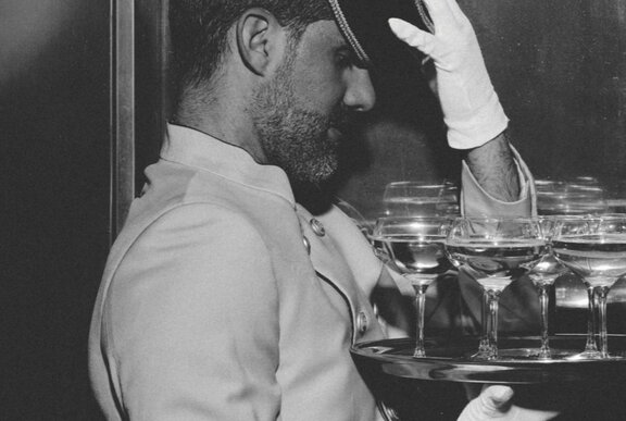 A black and white photo of a formally-dressed waiter with white gloves holding a tray of wide-mouth champagne glasses.