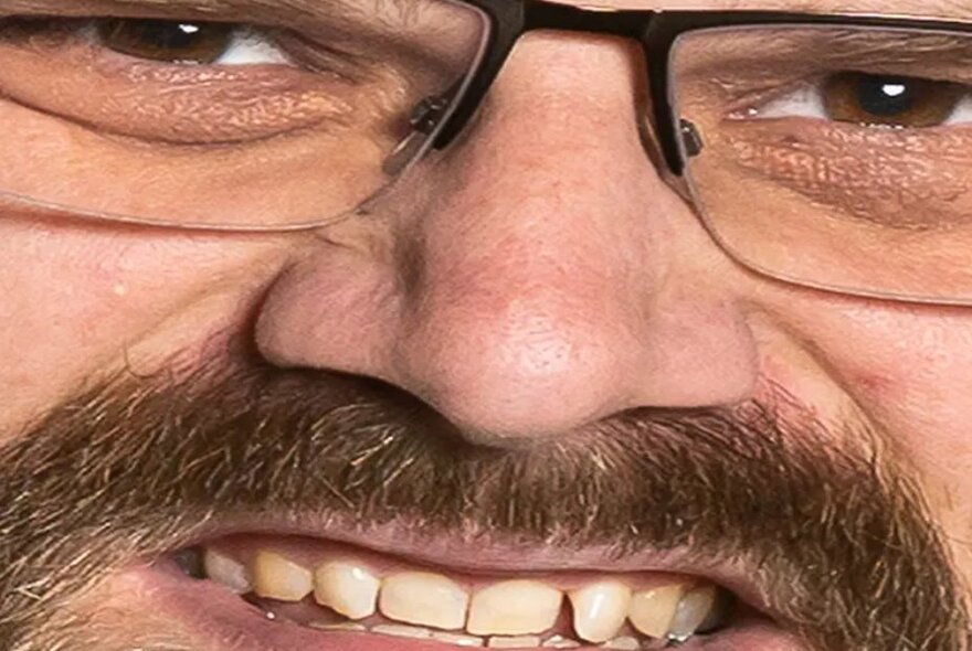 Extreme close-up of a man's face with moustache and glasses, gritted teeth.