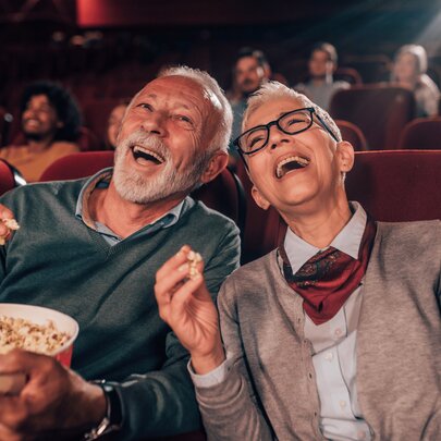 An older couple laughing while eating popcorn inside a cinema.