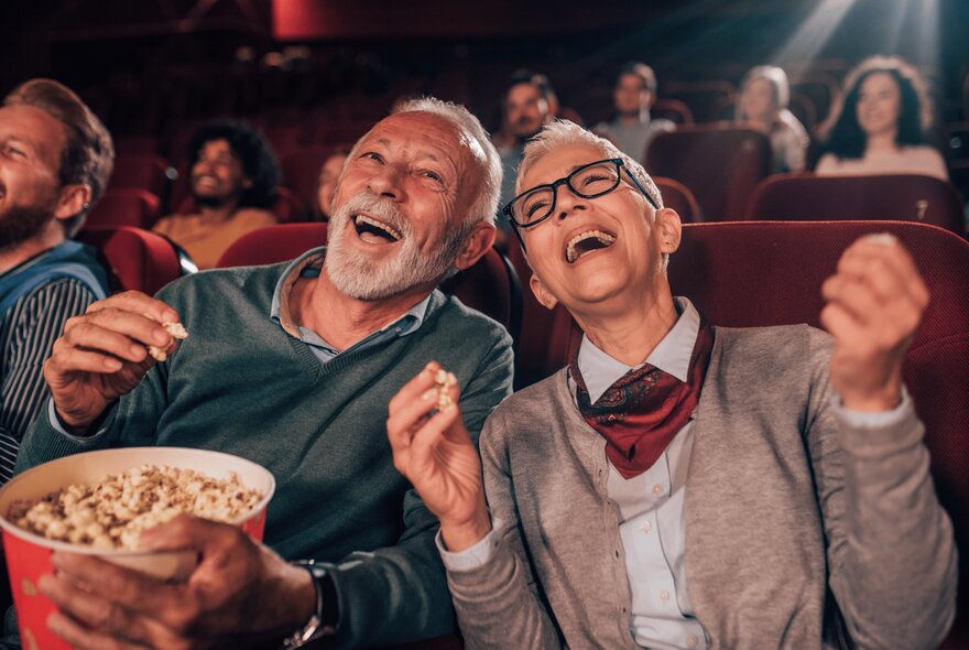 An older couple laughing while eating popcorn inside a cinema.