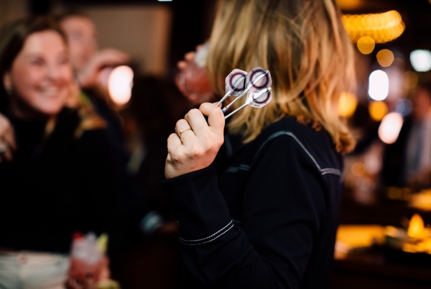 Person, from behind, with long sandy-blonde hair, holding three darts in left hand, cocktail in right.