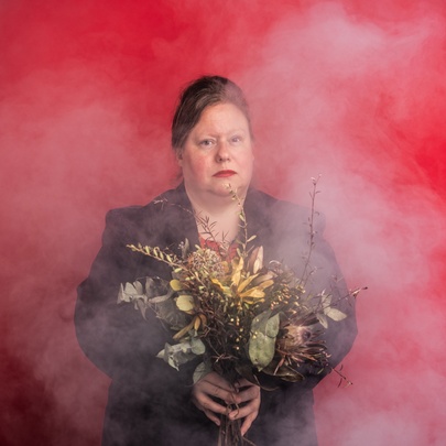 Actor Juliet Sironi holding a bunch of dried flowers with smoke all around her against a red background. 