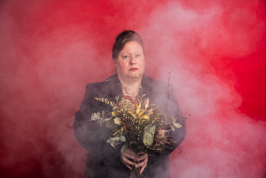 Actor Juliet Sironi holding a bunch of dried flowers with smoke all around her against a red background. 