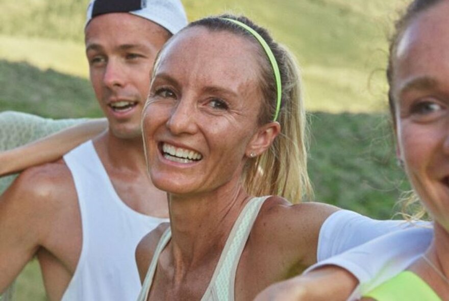 Woman with high ponytail and thin headband, wearing sporty tank top, sweaty and smiling, with two other people at side.
