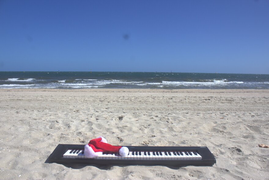 A digital piano keyboard on a beach with a red Christmas hat placed on top of it.