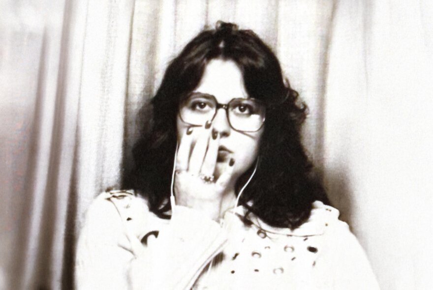 A black and white photograph of a person with dark, wavy hair and glasses, looking directly at the camera with their hand covering part of their mouth.