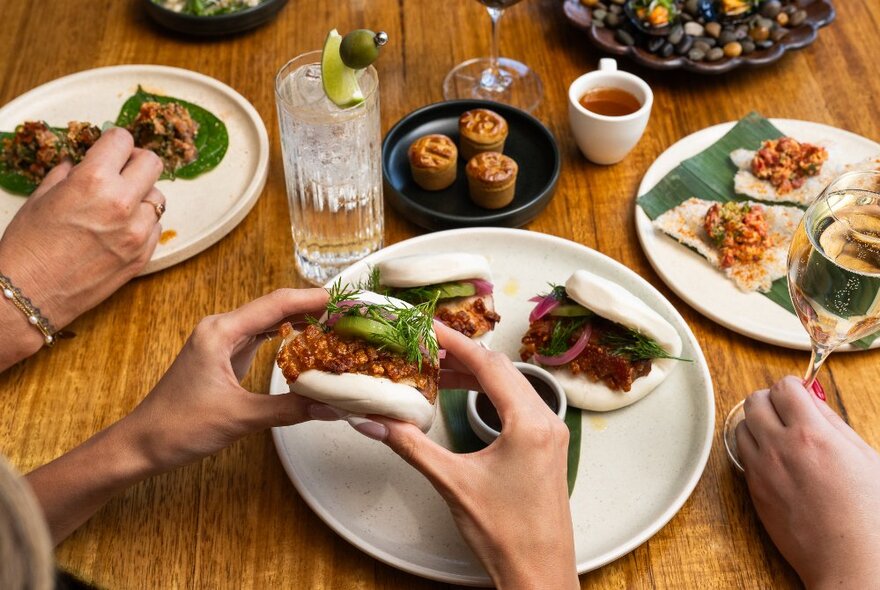 Hands holding bite-sized portions of food over white plates of small snacks, on a timber dining table.