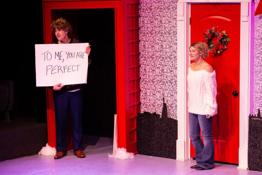 Actors recreating a scene from the film Love Actually, a man holding a sign reading 'To me, you are perfect', a woman standing in front of a front door decorated with a Christmas wreath.