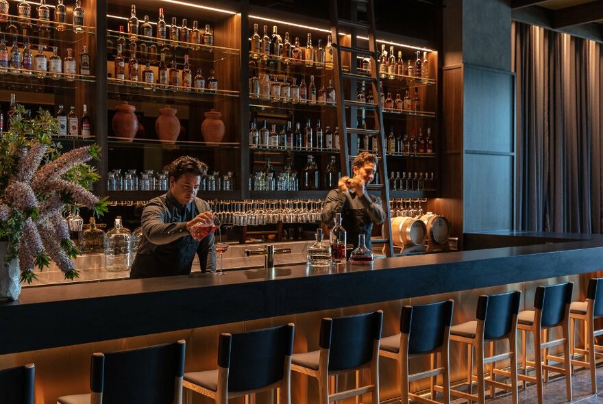 Two bartenders mixing drinks behind a long bar, the wall behind them filled with bottles.