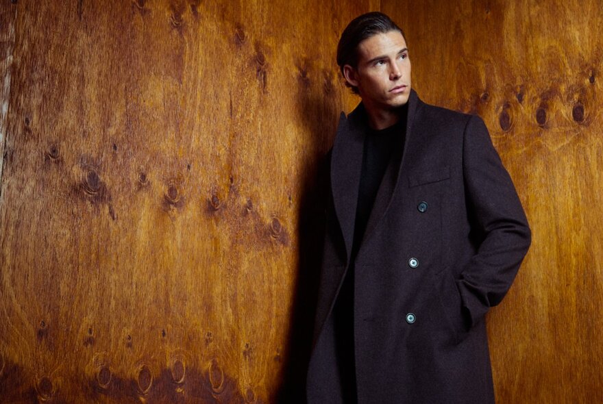 A male model wearing a tailored dark coat and leaning against a wood grain wall.