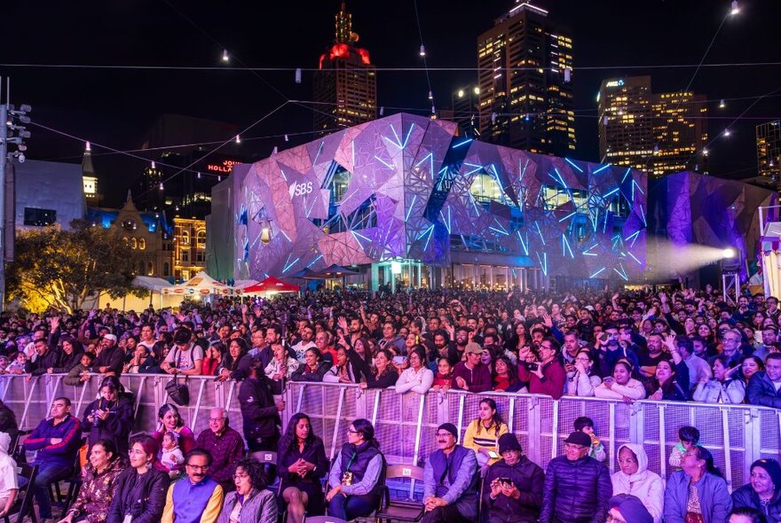 View of a large crowd of people at the Diwali Festival celebrations in the outdoor plaza at Fed Square, all facing the main stage, night time, with city buildings in the background.