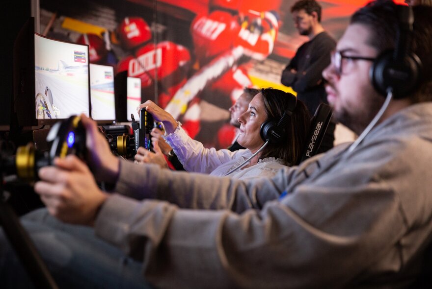 People in front of screens on racing driver simulators with headsets on and steering wheels in hard. 