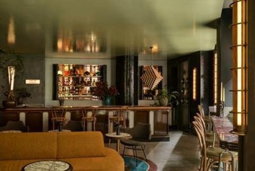 A stylish, warmly lit interior view of a bar, featuring a long wooden bar, a mustard-yellow sofa, and a tiled floor.