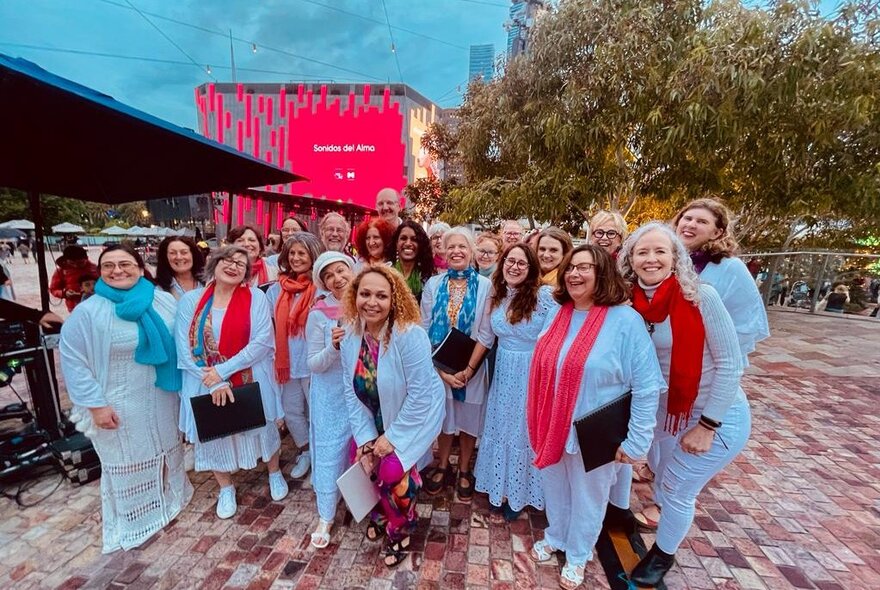A group of smiling people, wearing white outfits, some with red scarfs, standing close together in an open outdoor space.