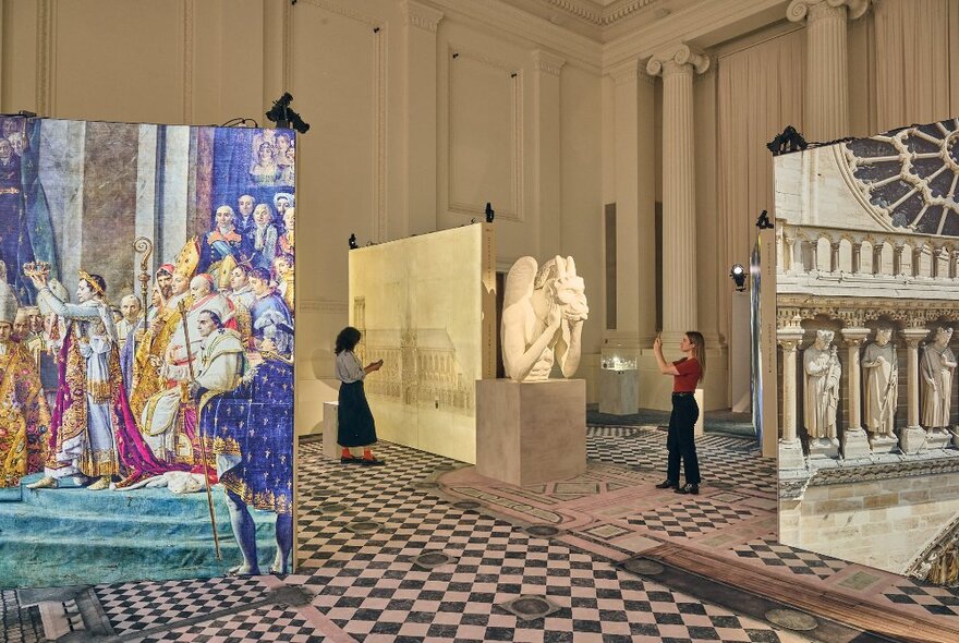 A large print of a colourful painting depicting the crowning of a monarch, inside a large room with checked carpet and marble pillars, patrons looking at other works in the background.