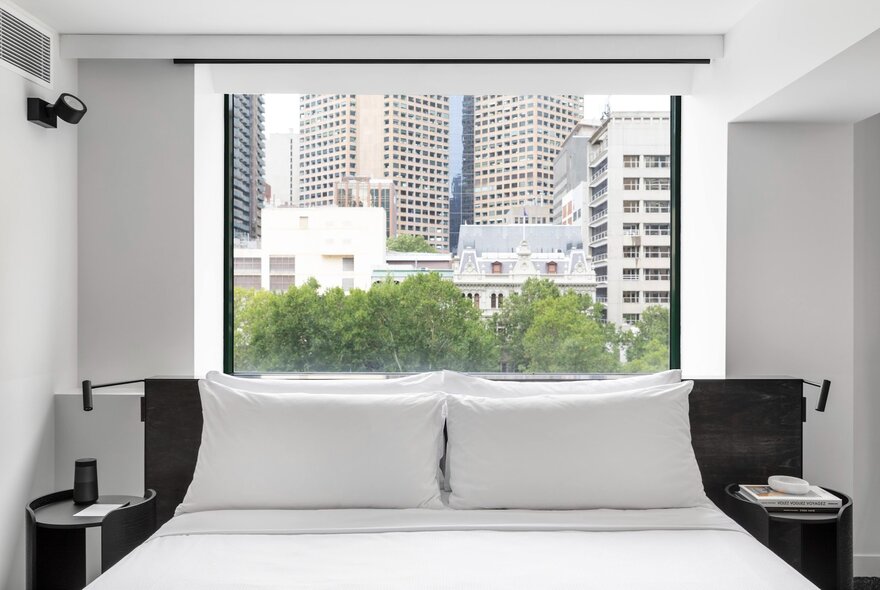 A room at the Lancemore Crossley St hotel in Melbourne, showing a king size bed positioned below a large window with views of tree tops and city buildings.