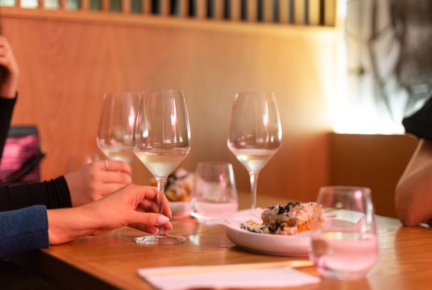 Two different hands holding two wine glasses, resting on a restaurant's dining table, a plate of food and napkins also on the table.