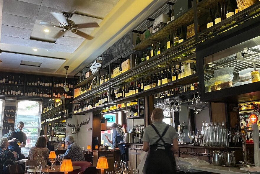 Staff and diners in a lamplit bar-restaurant with shelves of bottles and glasses.