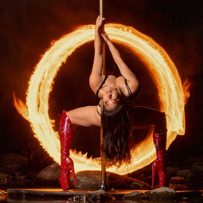 A performer with thigh-high red boots on a pole doing a deep backbend, framed by a blazing ring of fire in a dark setting.