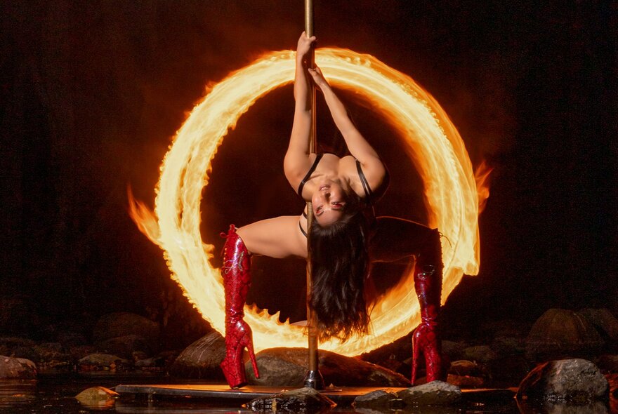 A performer with thigh-high red boots on a pole doing a deep backbend, framed by a blazing ring of fire in a dark setting.