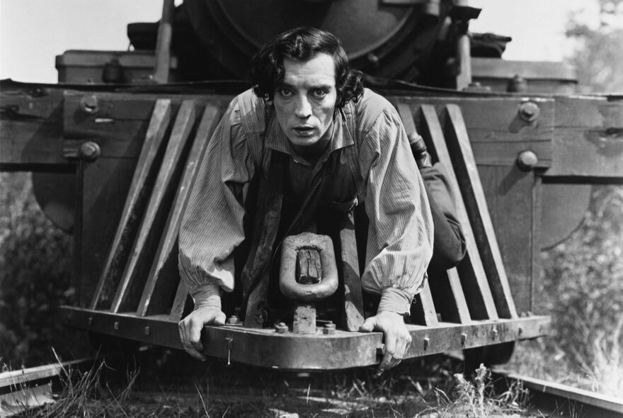 A black and white still from the silent film, The General, showing a young man hanging on to the pilot (the metal grill) at the front of a steam train. 