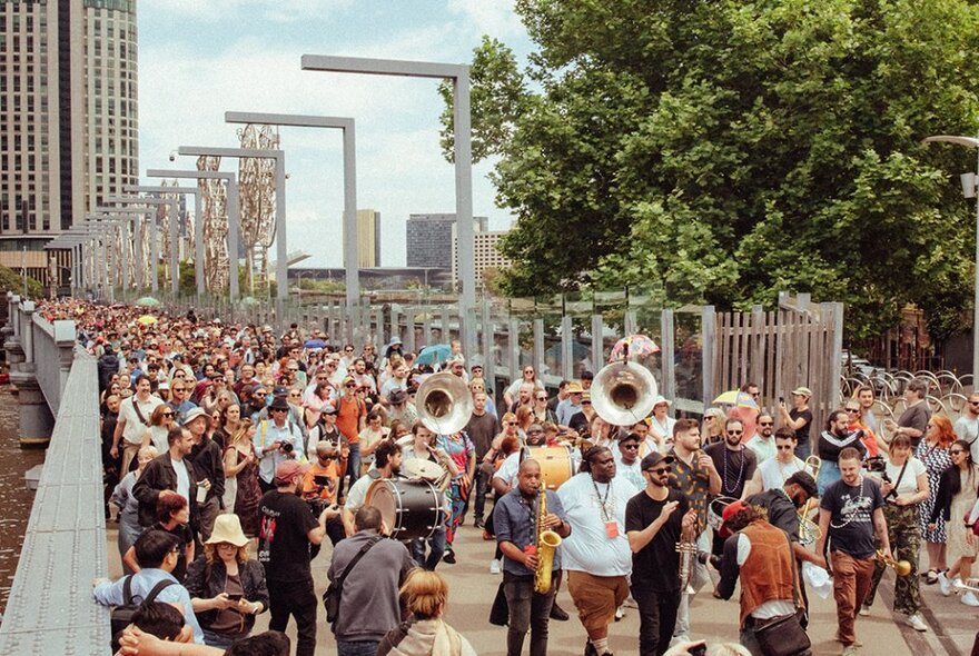 A large parade of people following a brass band across a bridge in a city, in a walking musical street party.