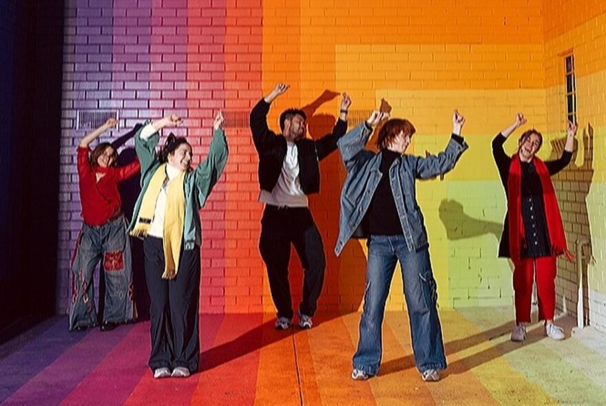 Five dancers each with their arms in the air, standing on a rainbow-coloured floor in front of a rainbow-coloured painted wall.
