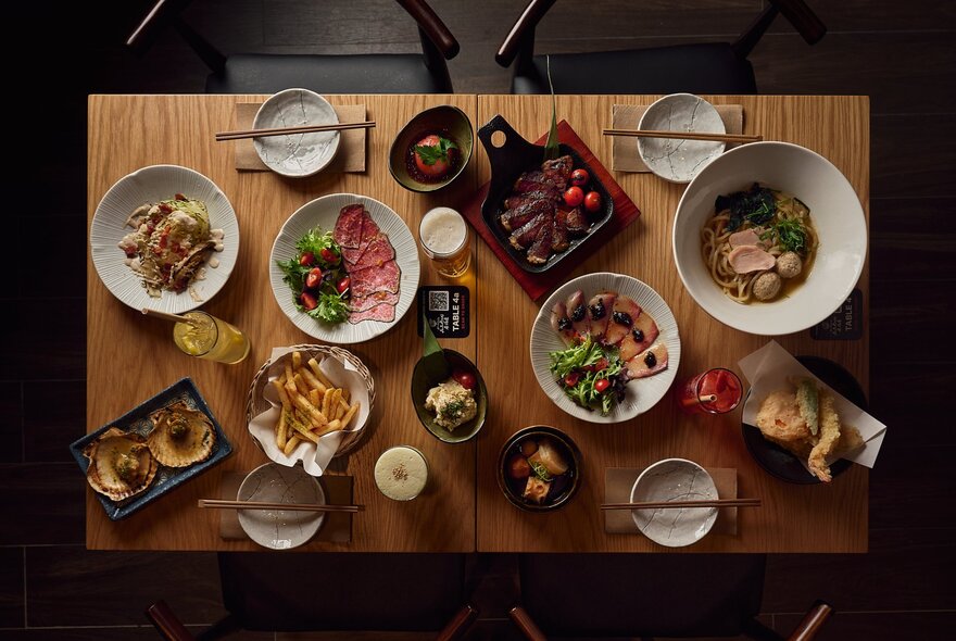 Overhead view of several Asian dishes of food on a wooden restaurant table with chopsticks.