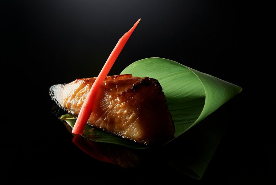 A slice of blackened salmon resting on a rolled banana leaf with a red garnish; black background.