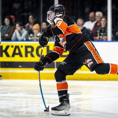 An ice hockey player in black and orange skating on an ice hockey rink; referee in the background.