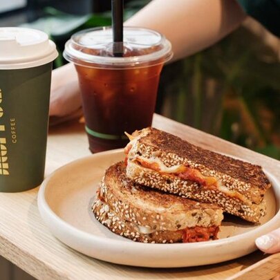 A person's hands holding a wooden tray with a green takeaway coffee cup, an iced coffee in a clear cup and a plate with a toasted sandwich.