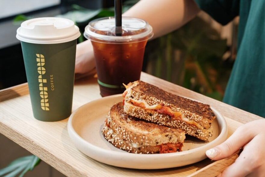 A person's hands holding a wooden tray with a green takeaway coffee cup, an iced coffee in a clear cup and a plate with a toasted sandwich.