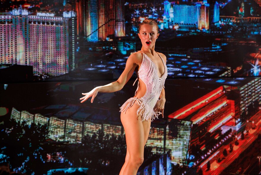 A dancer in a beaded costume dancing on stage in front of a backdrop that is an image of a city at night with buildings and bright lights.