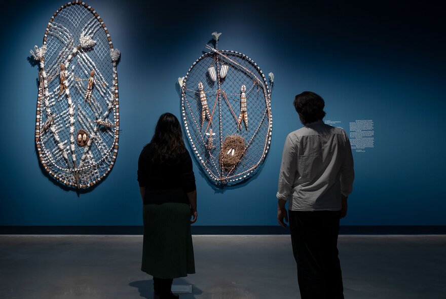 People viewing Indigenous craft artworks hanging on a blue wall in a gallery.