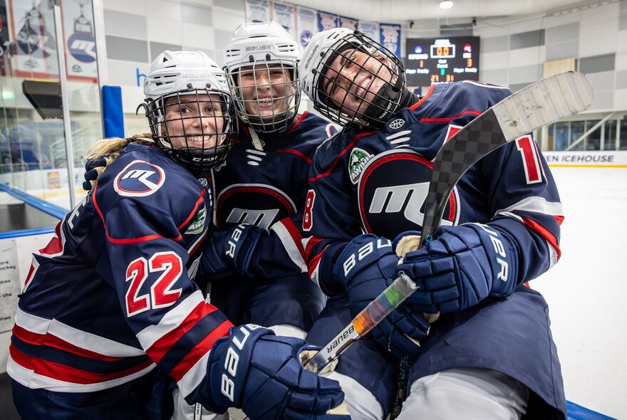 Three smiling female ice hockey players in a huddle wearing padded team outfits and helmets.