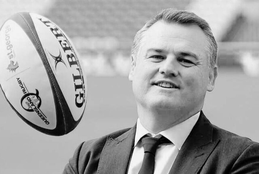 Black and white portrait photo of rugby great, Nick Stiles, wearing a suit and throwing a rugby ball into the air.
