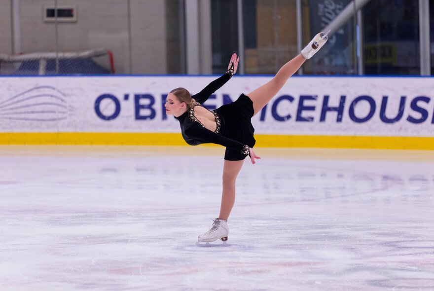 An ice skater in a black dress gliding with her leg in the air on the O'Brien Icehouse rink. 