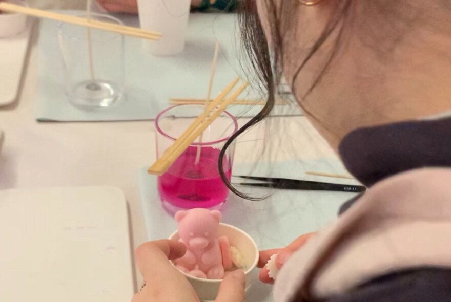 Looking over a person's shoulder to see them making a candle with a small pink teddy bear on top. 