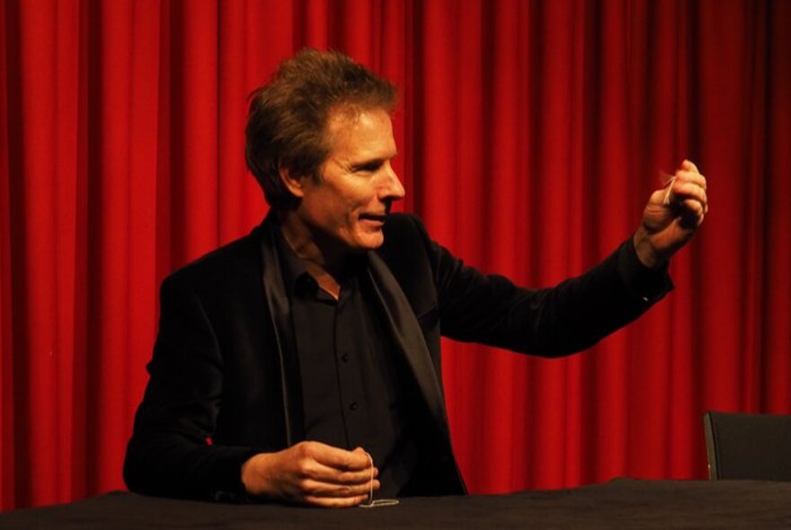 Magician Tim Ellis performing a close-up illusion at a table in front of a red curtain.