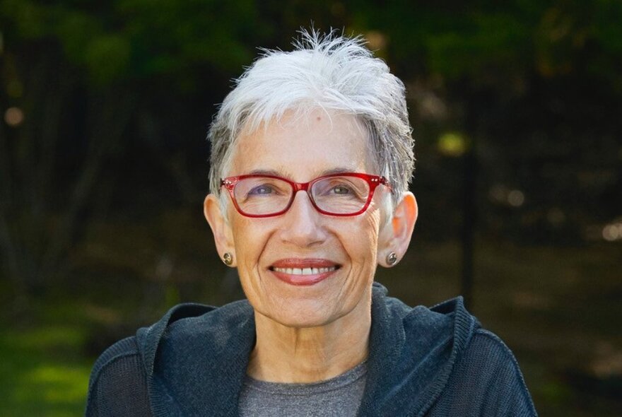 Author Dava Sobel, a middle-aged woman with short white hair, red rimmed glasses and a smile on her face, staring directly at the viewer, in an outdoor location.