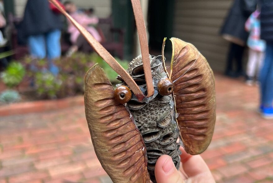 A craft object made from seed pods, sticks, and dried foliage, being held by a small hand.